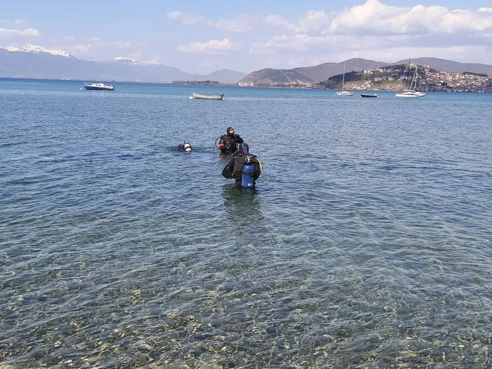 Two scuba divers in shallow water at Kaj Divono beach with boats and the city of Ohrid in the background