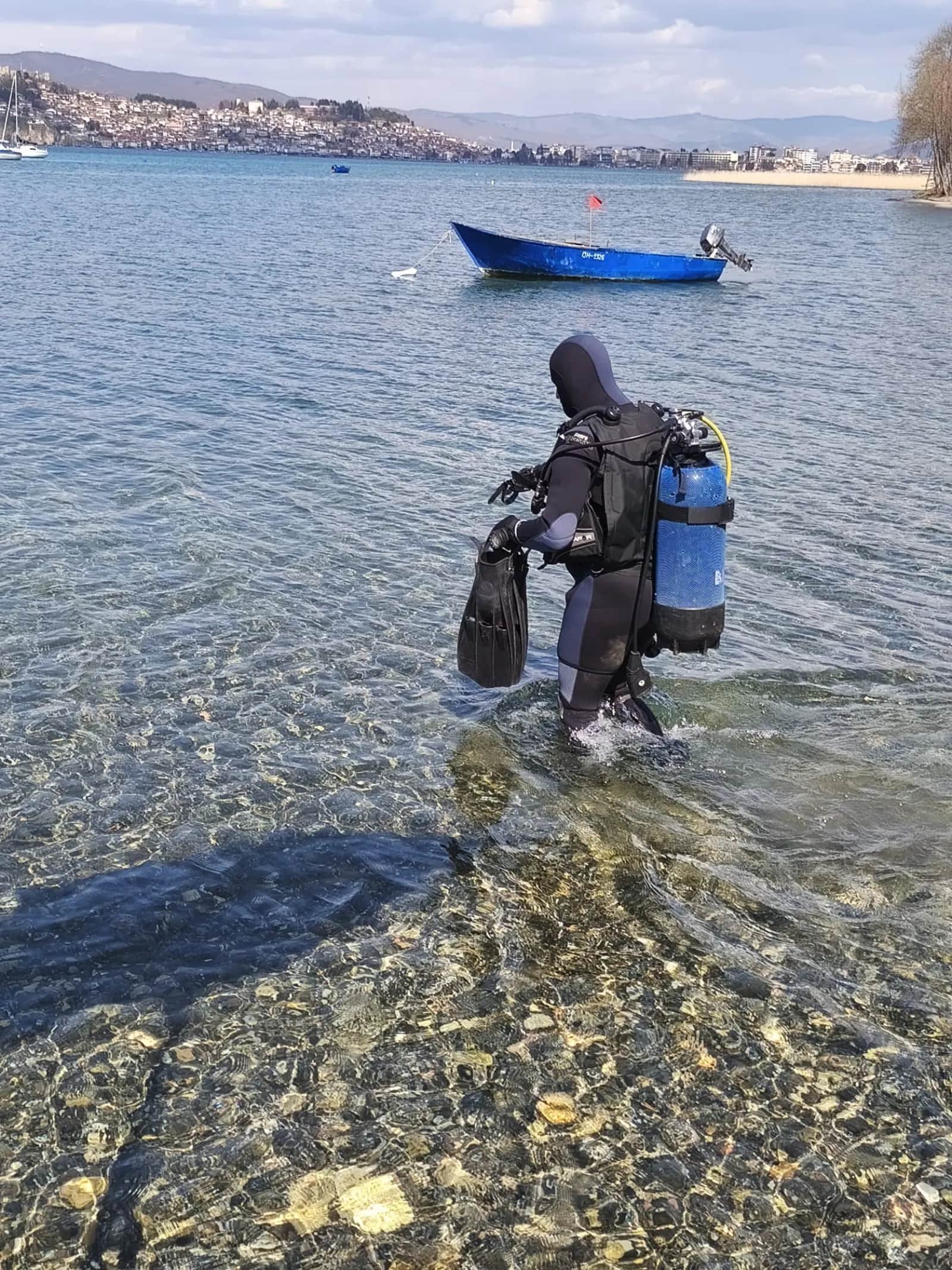A scuba diver walking out of Lake Ohrid carrying full diving gear, with the Old Town of Ohrid in view
